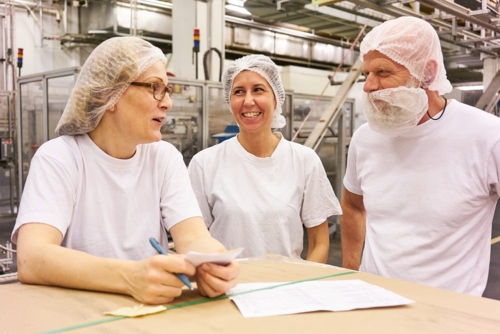 tre estrellaanställda i produktion iklädda vit tshirt och hårnät. de skarttar tillsammans.